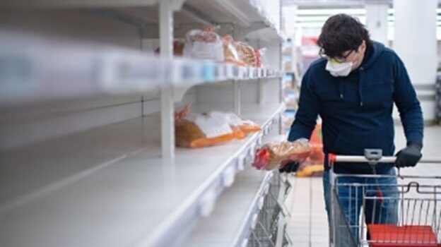 A man with a mask picking up a loaf of bread from near-empty supermarket shelves