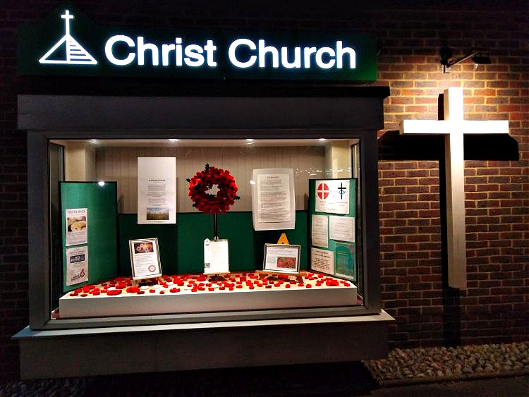 The end of the Christ Church building at night showing the illuminated Christ Church sign, a white cross and a lit Remembrance Sunday window display with poppies and a poppy wreath