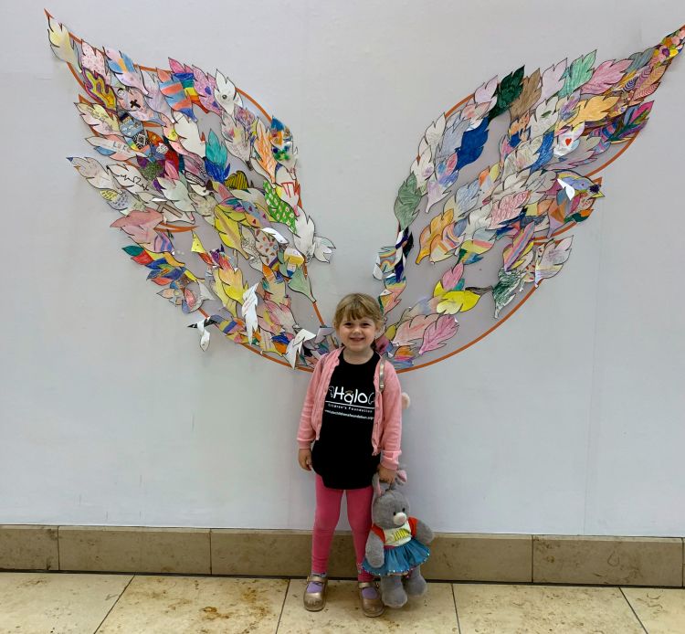 A little girl wearing a 'Halo Children's Foundation' T-shirt standing in front of a wall with angel wings