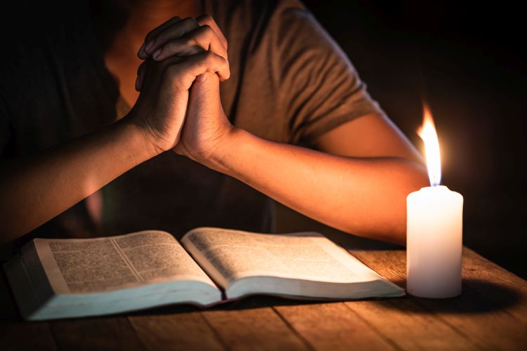 A man sitting at a table praying with an open Bible and a lit candle in front of him