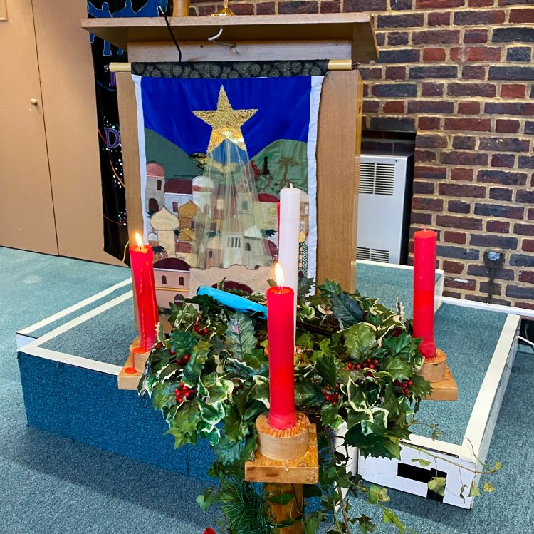 An Advent wreath with five candles in front of the lectern in the church which has a hanging on the front depicting the Nativity scene