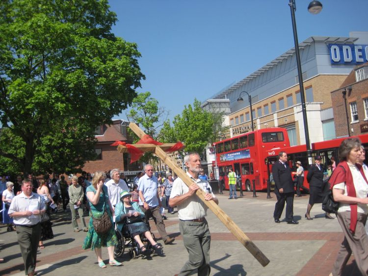 A man carrying a wooden cross through Uxbridge town centre with a crowd following