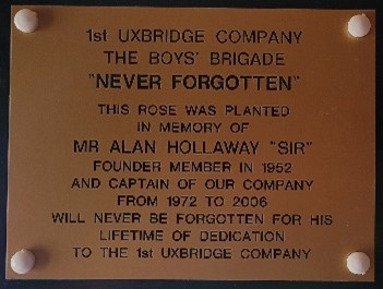 A plaque reading "1st Uxbridge Company The Boys' Brigade. 'Never Forgotten'. This rose was planted in memory of Mr Alan Hollaway "Sir" founder member in 1952 and Captain of our Company from 1972 - 2006. Will never be forgotten for his lifetime of dedication to the 1st Uxbridge Company."