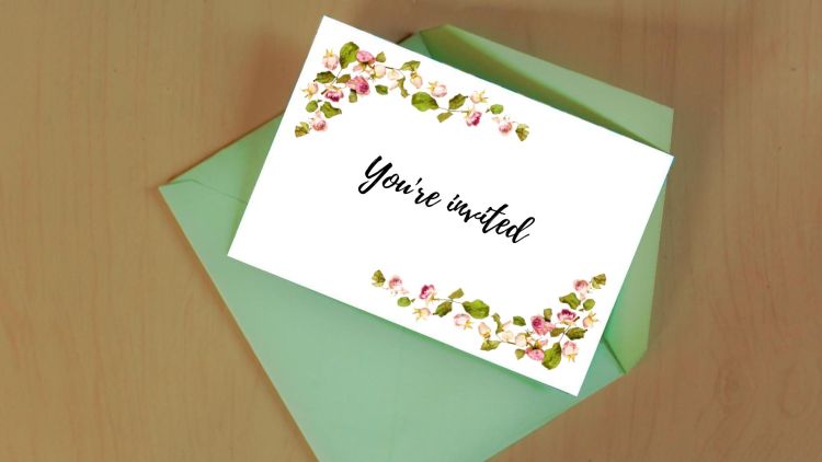 A card with a flowery border and the words 'You're invited' on top of a green envelope on a wooden surface