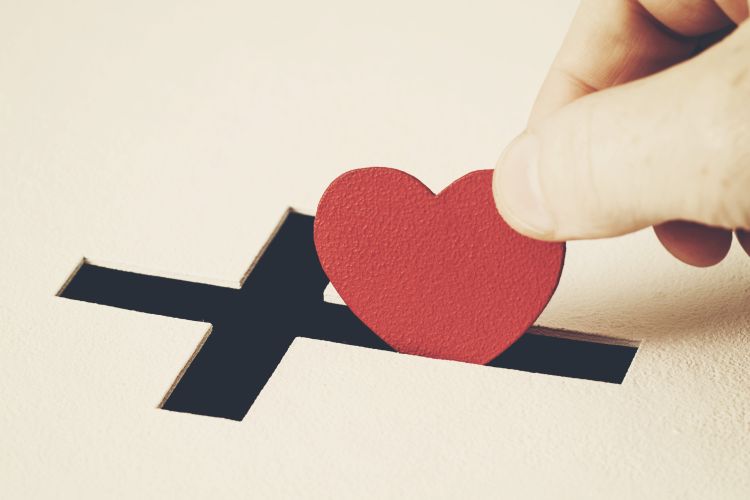 Red heart symbol is put by person's hand into slot of white donation box, slot is shaped like Christian cross. 