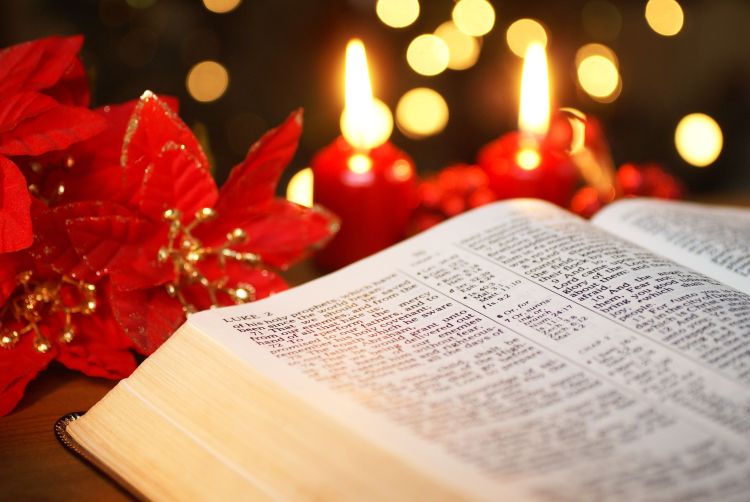 An open Bible with poinsettia flowers and red candles on the table behind it