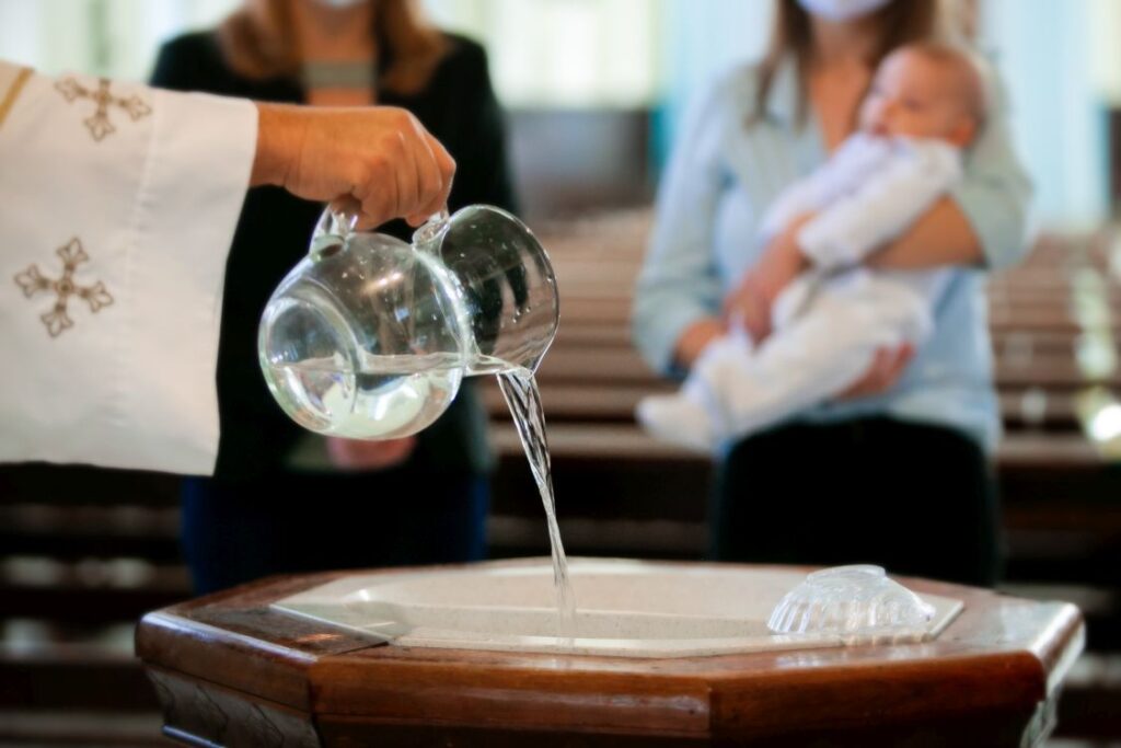 A priest pouring holy water into the baptismal font with parents and a child seen in soft focus in the background
