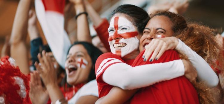 English female soccer fans with England flag painted on their faces hugging each other after their team's victory. English female spectators in football stadium celebrating their team's victory.