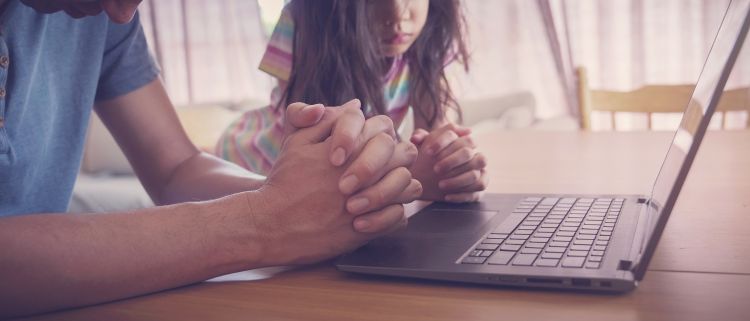 An adult and child praying in front of an open laptop
