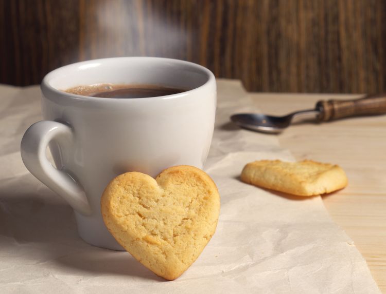 A steaming cup of coffee with a heart shaped biscuit leaning against the cup