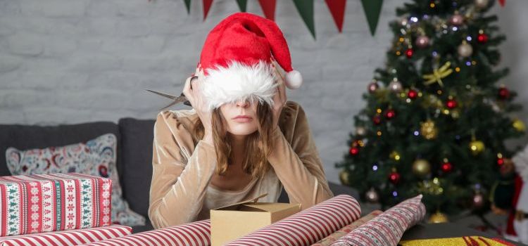 A woman with a Santa hat pulled down over her eyes and her head in her hands. There is a Christmas tree behind her and a table in front of her, covered in wrapping paper and boxes