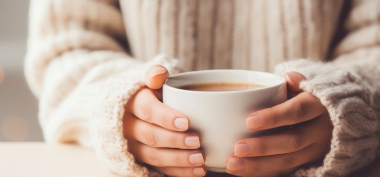 A person in a cream knitted jumper with their hands wrapped around a mug of tea.
