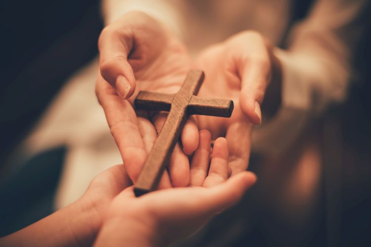 Two pairs of hands touching with palms upwards. A wooden cross lies across the two pairs of hands